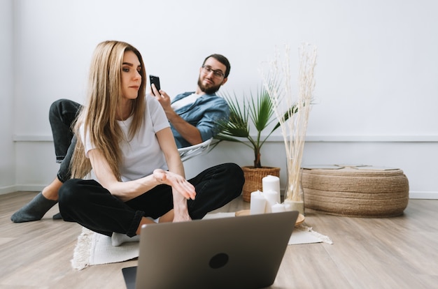 Jeune couple utilisant un ordinateur portable, parcourant les informations et regardant l'écran du portable dans le salon moderne.