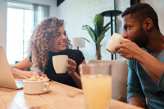 Jeune couple utilisant un ordinateur portable tout en prenant le petit déjeuner ensemble à la maison.