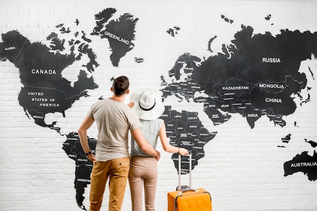Jeune couple de voyageurs debout près de la grande carte du monde en arrière-plan choisissant un lieu pour des vacances d'été