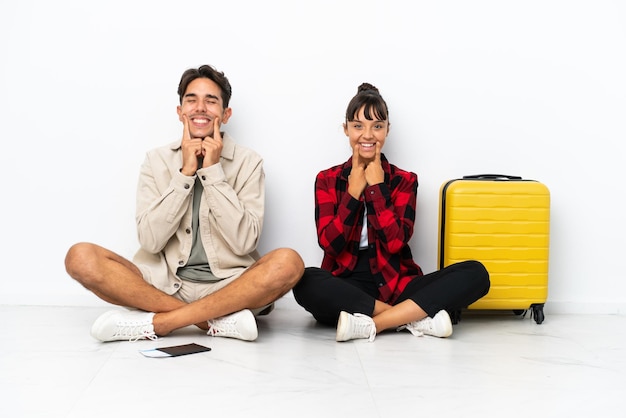 Jeune couple de voyageurs métis assis sur le sol isolé sur fond blanc souriant avec une expression heureuse et agréable