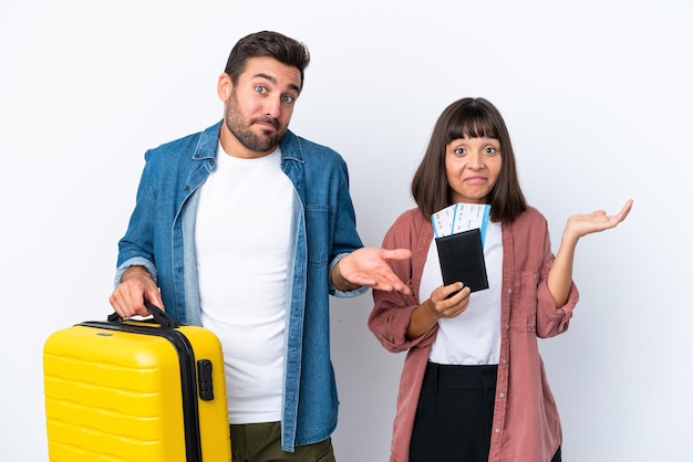 Jeune couple de voyageurs tenant une valise et un passeport isolé sur fond blanc ayant des doutes tout en levant les mains et les épaules