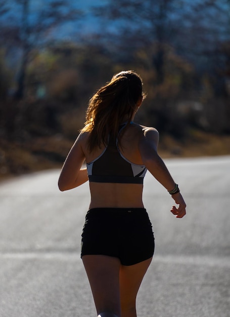 Une jeune coureuse en forme sur le dos portant un short serré et un t-shirt sportif serré courant avec style sur un sentier d'asphalte éclairé par les rayons du soleil de l'aube dans un parc naturel