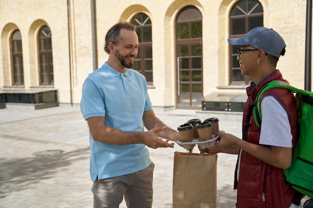 Photo jeune courrier masculin asiatique avec sac thermo donnant quatre tasses de café