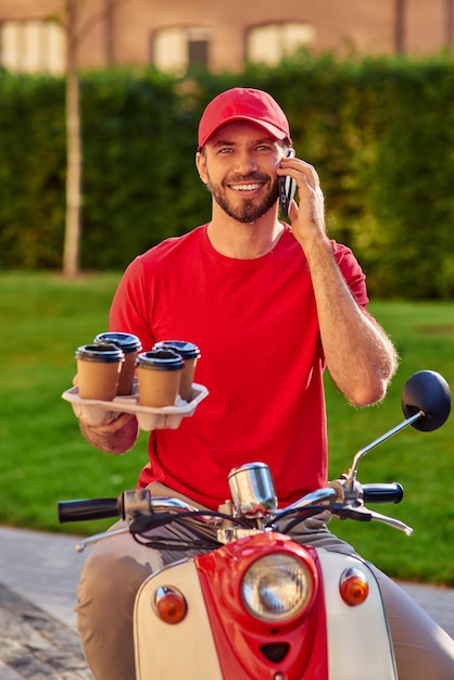 Jeune courrier masculin caucasien parlant par smartphone et souriant tout en livrant quatre tasses de café par