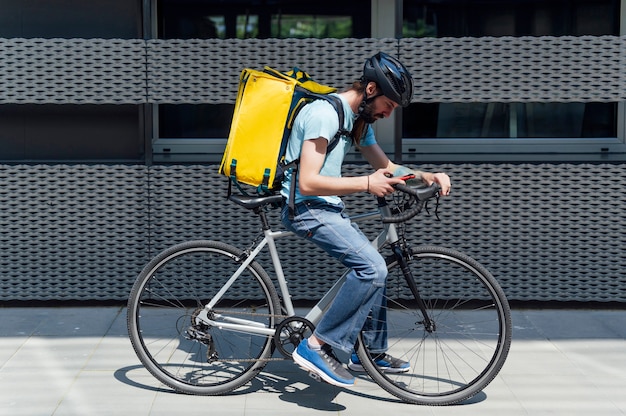 Jeune coursier barbu livrant de la nourriture sur un vélo, vérifiant la commande avec un téléphone intelligent. Concept de service de livraison