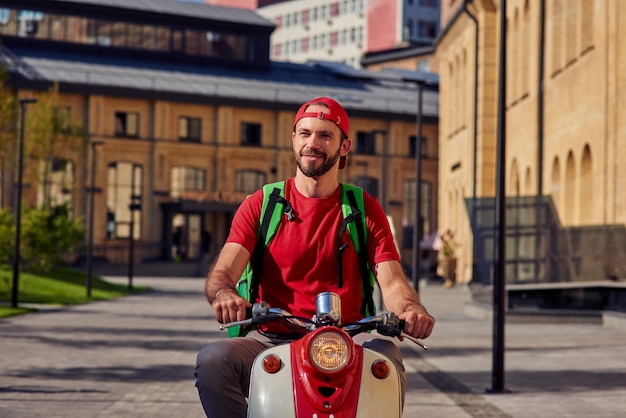 Jeune coursier masculin caucasien gai avec sac de réfrigérateur regardant de côté tout en conduisant un scooter le long d'une rue ensoleillée. Concept de livraison de nourriture