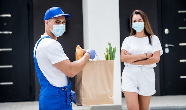 Un jeune coursier portant un masque de protection et des gants livre des marchandises à une jeune femme pendant la quarantaine