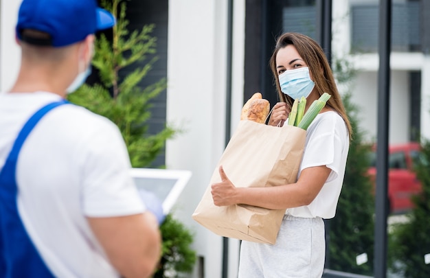 Un jeune coursier portant un masque de protection et des gants livre des marchandises à une jeune femme pendant la quarantaine