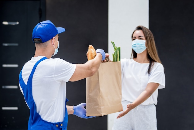 Un jeune coursier portant un masque de protection et des gants livre des marchandises à une jeune femme pendant la quarantaine