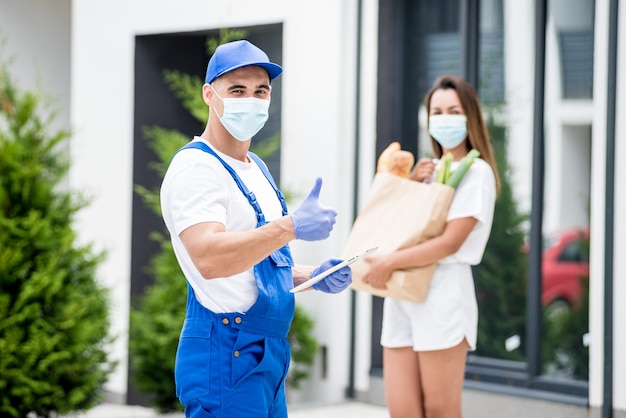 Un jeune coursier portant un masque de protection et des gants livre des marchandises à une jeune femme pendant la quarantaine