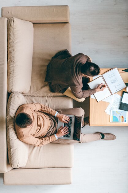 Jeune courtier prenant des notes dans le bloc-notes lors de la planification des travaux pendant que son collègue avec un ordinateur portable travaille sur le projet