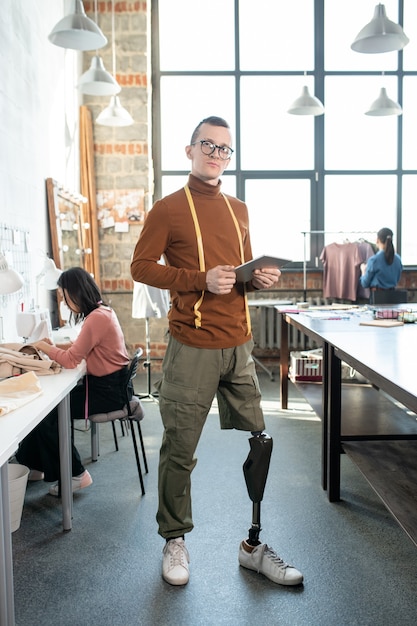 Jeune créateur de mode ou tailleur avec jambe prothétique utilisant une tablette par lieu de travail contre des collègues féminines à l'intérieur d'un grand atelier