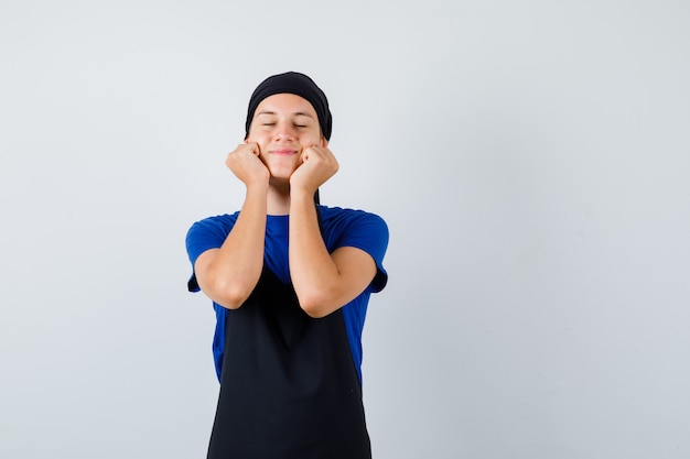 Jeune cuisinier adolescent en t-shirt, tablier penché les joues sur les mains, fermant les yeux et l'air joyeux, vue de face.