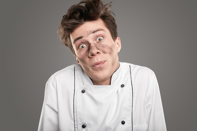 Jeune cuisinier drôle avec de la suie sur le visage, haussant les épaules et regardant la caméra tout en s'excusant d'avoir commis une erreur sur fond gris