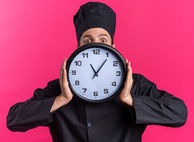 Jeune Cuisinier Masculin Blond Concerné En Uniforme De Chef Et Casquette Tenant Une Horloge Regardant La Caméra Par Derrière Isolé Sur Un Mur Rose