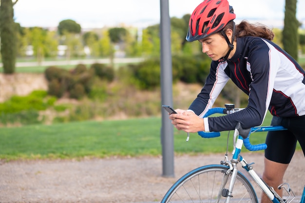 Jeune cycliste masculin fatigué, vêtu de sport et casque, utilisant un smartphone tout en se détendant après avoir fait du vélo dans le parc