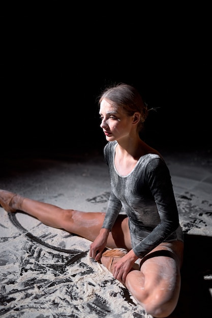 Jeune danseuse de ballet fatiguée en body assise sur le sol avec de la farine tout en se reposant après un entraînement épuisant ou une performance en studio sur fond noir, une ballerine talentueuse est assise détendue.