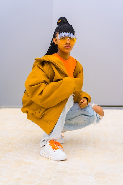 Une jeune danseuse de trap avec des tresses. Fille ethnique noire avec T-shirt, lunettes de soleil orange et chasseur, Urban Posé Buck