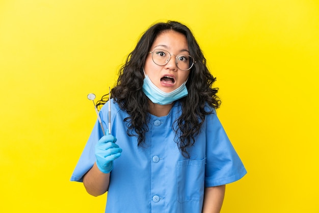 Jeune dentiste asiatique tenant des outils sur fond isolé avec une expression faciale surprise