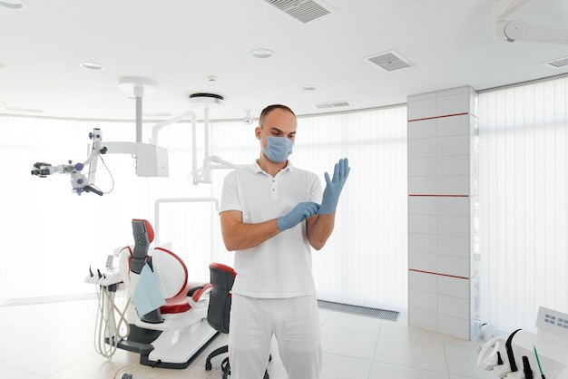 Un jeune dentiste dans un masque se tient près d'un fauteuil dentaire rouge et sourit dans la dentisterie blanche moderne Dentisterie et prothèses modernes