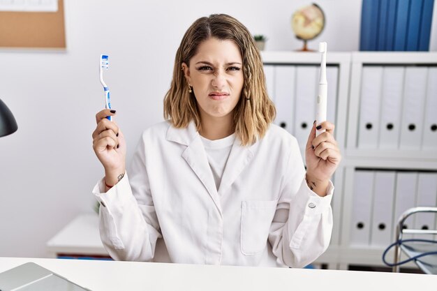 Jeune dentiste hispanique tenant une brosse à dents électrique et une brosse aux dents à la clinique expression désemparée et confuse concept de doute