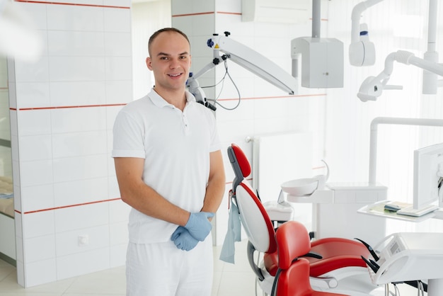 Un jeune dentiste se tient près d'un fauteuil dentaire rouge et sourit dans la dentisterie blanche moderne Traitement et prévention des caries chez les jeunes Dentisterie et prothèses modernes