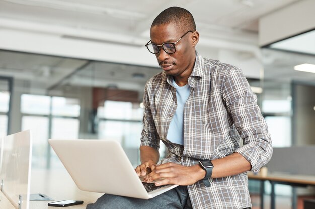 Jeune designer africain à lunettes en tapant sur un ordinateur portable, il travaille en ligne au bureau