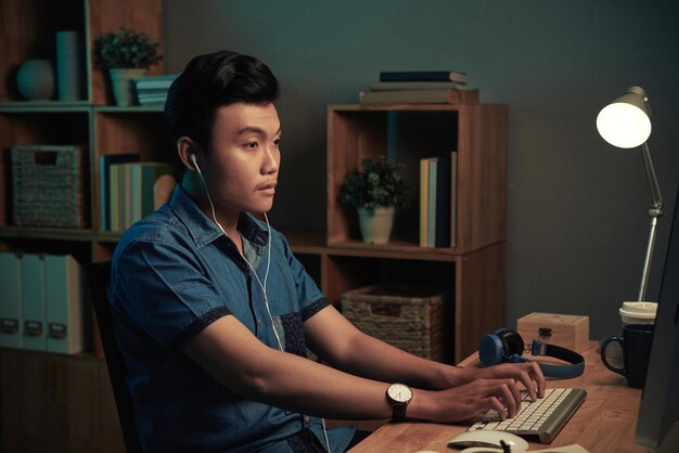 Jeune designer asiatique sérieux et concentré dans les écouteurs assis au bureau et tapant sur le clavier de l'ordinateur dans une pièce sombre