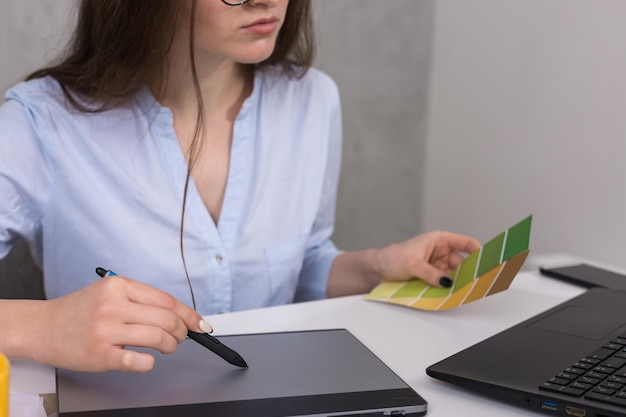 Une jeune designer assise à une table dessine sur une tablette. Effectue des calculs commerciaux sur l'ordinateur.