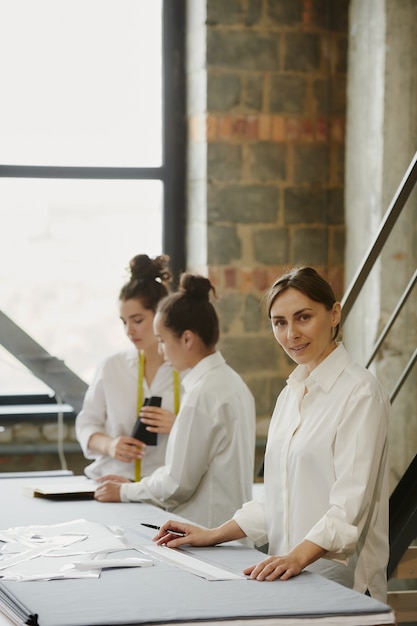 Jeune designer confiant de vêtements vous regarde en se tenant debout par table sur le mur de deux collègues discutant de nouveaux croquis