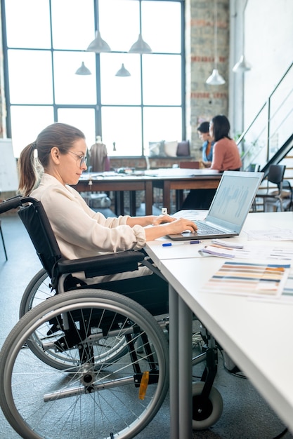 Jeune designer créatif en fauteuil roulant utilisant un ordinateur portable tout en dessinant des croquis pour une nouvelle collection saisonnière contre deux collègues en atelier