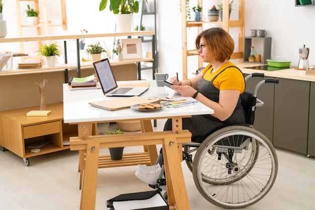 Jeune designer féminin sérieux en fauteuil roulant, dessin de croquis de nouveaux éléments technologiques avec l'aide de la tablette et du stylet à la maison