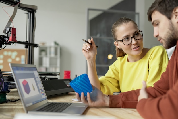 Jeune designer femme confiante parlant à son collègue avec un objet en plastique bleu imprimé sur une machine d'impression 3D