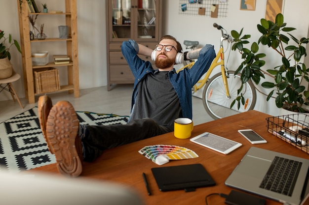 Jeune designer indépendant décontracté dans des vêtements décontractés et des écouteurs gardant ses jambes sur la table tout en écoutant de la musique devant le lieu de travail