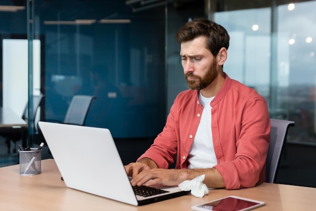 Un jeune designer masculin fatigué dans une chemise rouge travaille au bureau sur un ordinateur portable, il se sent mal, il est malade