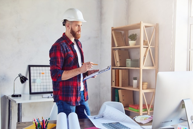 Jeune designer masculin prenant des notes dans les dessins et regardant par la fenêtre de son bureau. Concept d'ingénierie et d'architecture