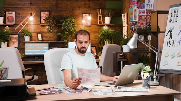 Jeune designer regardant des draps assis à son bureau avec un ordinateur portable devant. Espace de travail cosy et intelligent