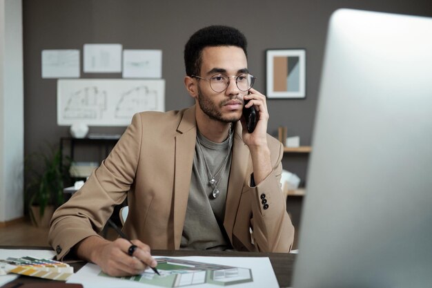 Jeune designer sérieux avec smartphone à l'oreille en regardant l'écran de l'ordinateur