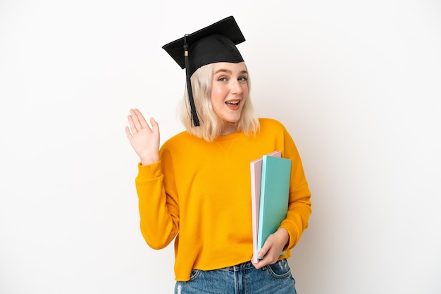 Jeune diplômé universitaire femme caucasienne isolé sur fond blanc saluant avec la main avec une expression heureuse