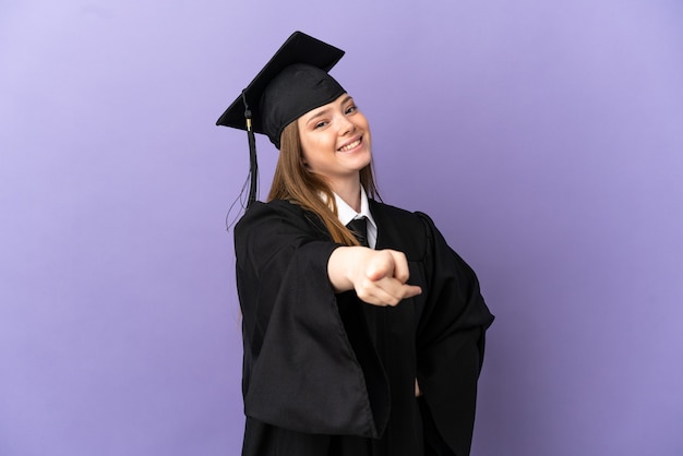 Jeune diplômé universitaire sur fond violet isolé pointant vers l'avant avec une expression heureuse