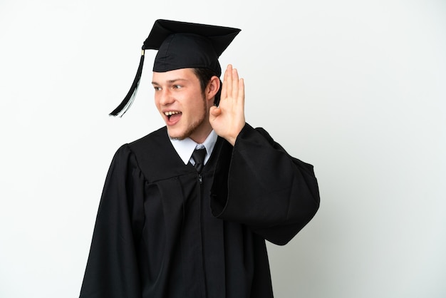 Jeune diplômé universitaire russe isolé sur fond blanc écoutant quelque chose en mettant la main sur l'oreille