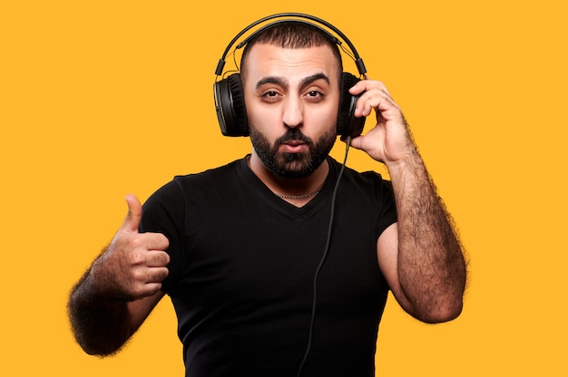 Jeune dj arabe avec barbe, écouter de la musique dans des écouteurs et montrant le pouce vers le haut jaune