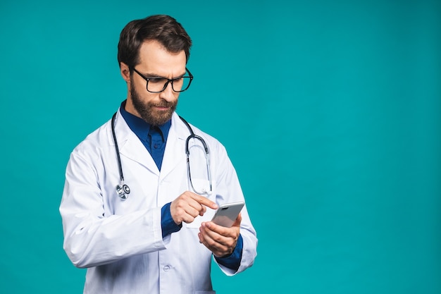 Jeune docteur textos sur un téléphone intelligent isolé sur fond bleu. Utilisation d'un téléphone portable.