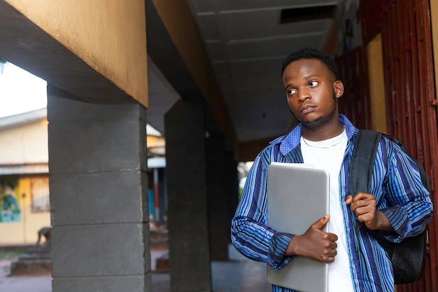 Jeune écolier adolescent africain triste avec sac à dos tenant un ordinateur portable