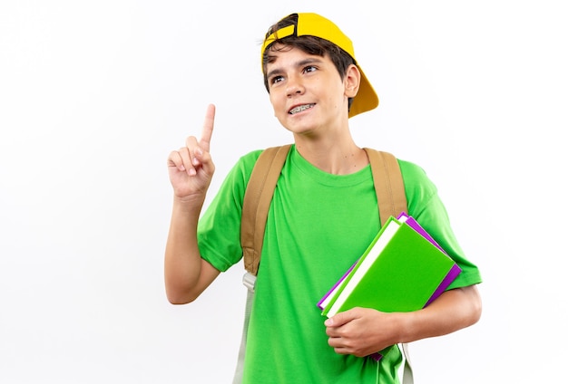 Un jeune écolier impressionné portant un sac à dos avec une casquette tenant des livres pointe vers le haut