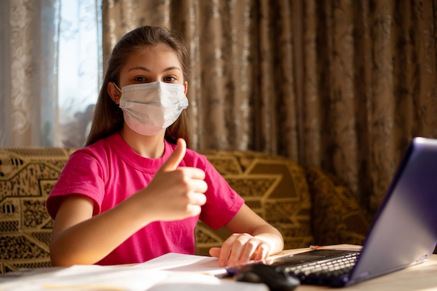 Jeune écolière en masque médical montrant les pouces vers le haut, heureuse d'avoir des cours à la maison, d'avoir une formation à distance et de ne pas aller à l'école. Concept de vie pendant la quarantaine des coronavirus