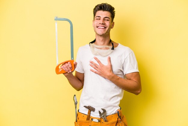 Jeune électricien caucasien isolé sur fond jaune rit bruyamment en gardant la main sur la poitrine.