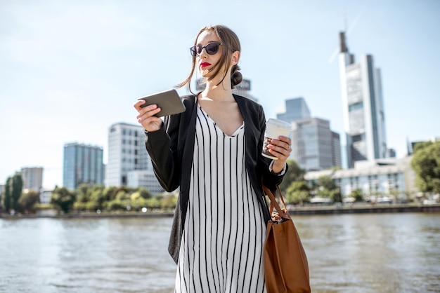 Jeune et élégante femme d'affaires marchant avec téléphone et café sur le quai avec des gratte-ciel dans la ville de Francfort