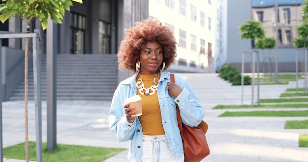 Jeune élégante Femme Afro-américaine Belle Bouclée Avec Sac Marchant Dans La Rue De La Ville, Buvant Du Café à Emporter Et Souriant Joyeusement. Femme Assez Heureuse Se Promener à L'extérieur Et Siroter Une Boisson Chaude.