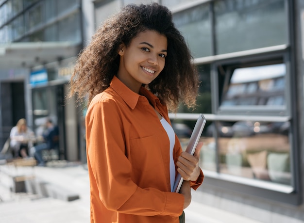 Jeune élégante femme afro-américaine tenant un ordinateur portable, marchant dans la rue